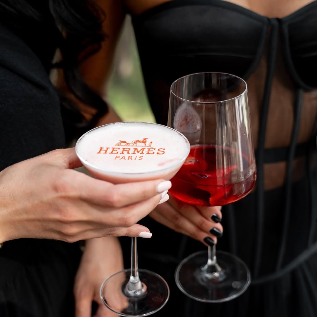 Cocktail printer με το λογότυπο Hermès στη φόα του κοκτέιλ δίπλα σε κόκκινο κρασί