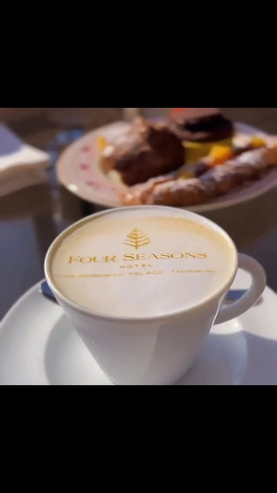 Latte art printer result: Four Seasons logo on coffee foam, with dessert plate in background